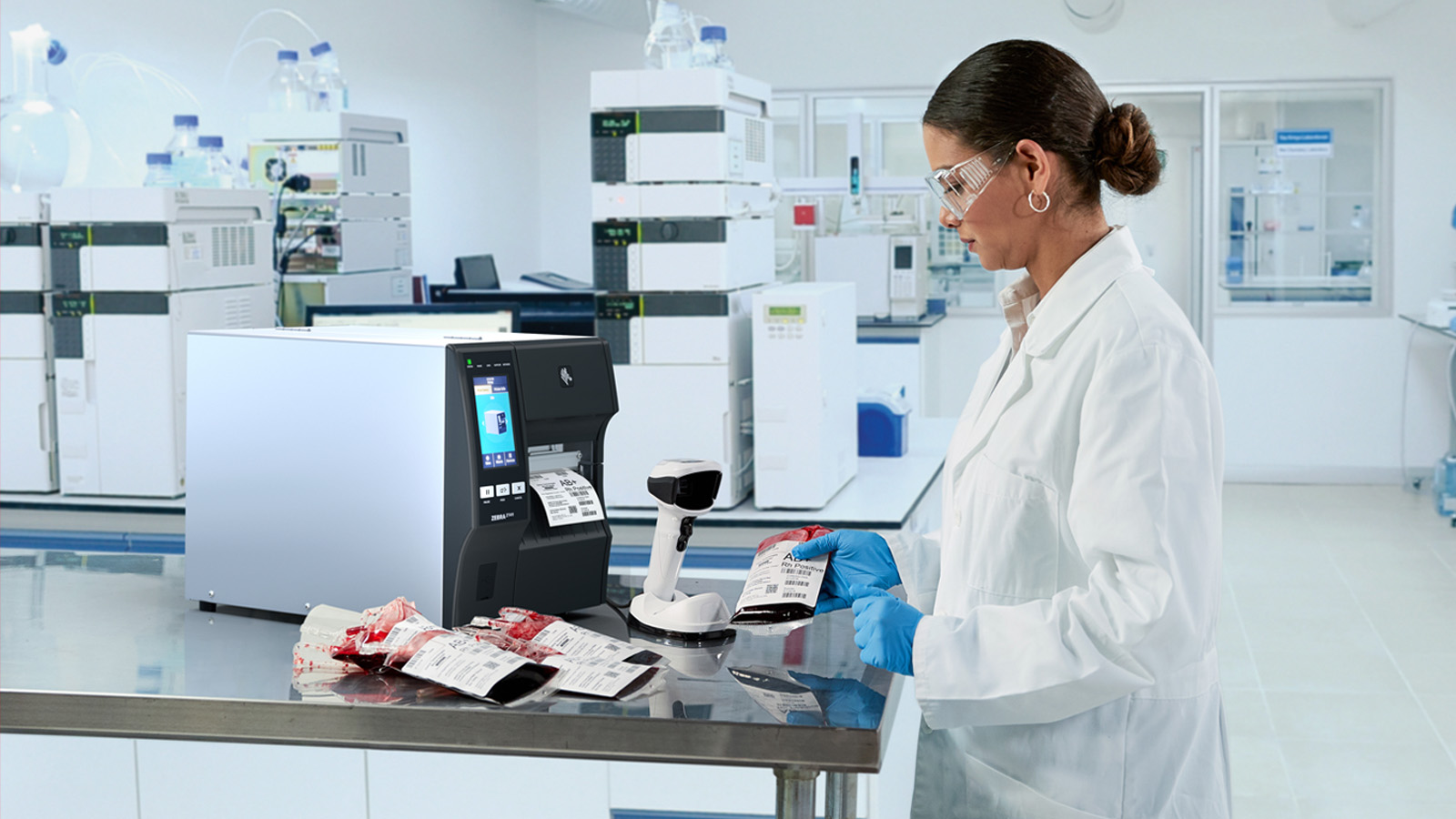 A healthcare professional is in a laboratory, holding a bag of blood to check the information on the label. She is standing next to a label printer and a handheld scanner.