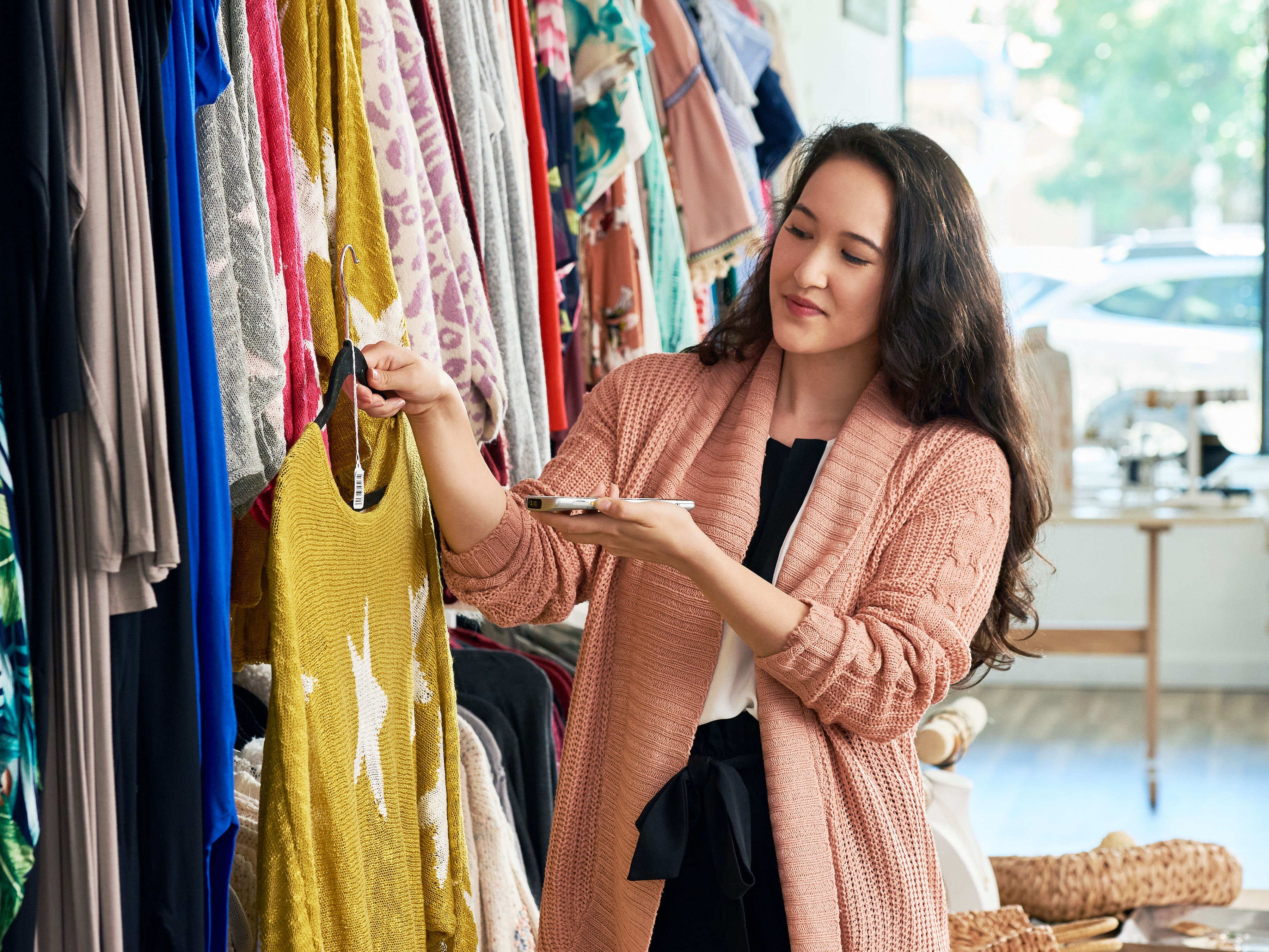 A woman in a clothing store holds a yellow sweater on a hanger while using a mobile computer to scan the garment’s tag, accessing its digital product passport (DPP).