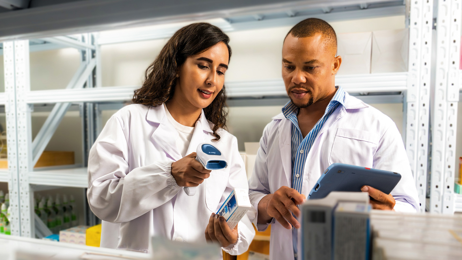 Two healthcare professionals in lab coats use health information technology (HIT) to review medication inventory; one uses a barcode scanner to scan a label, while the other consults a tablet for real-time updates. 
