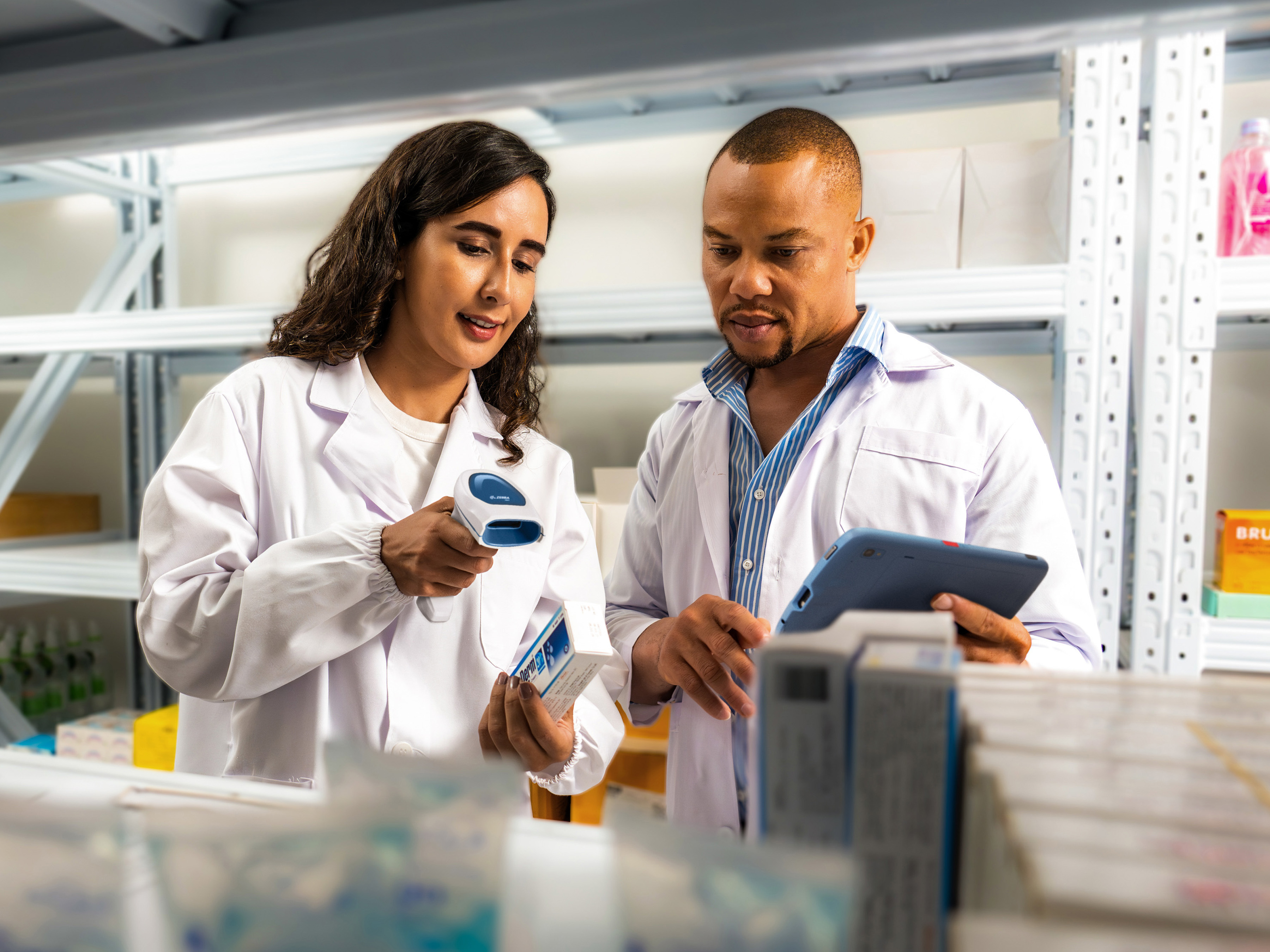 Two healthcare professionals in lab coats use health information technology to review medication inventory; one uses a barcode scanner to scan a box, while the other consults a tablet for real-time updates. 