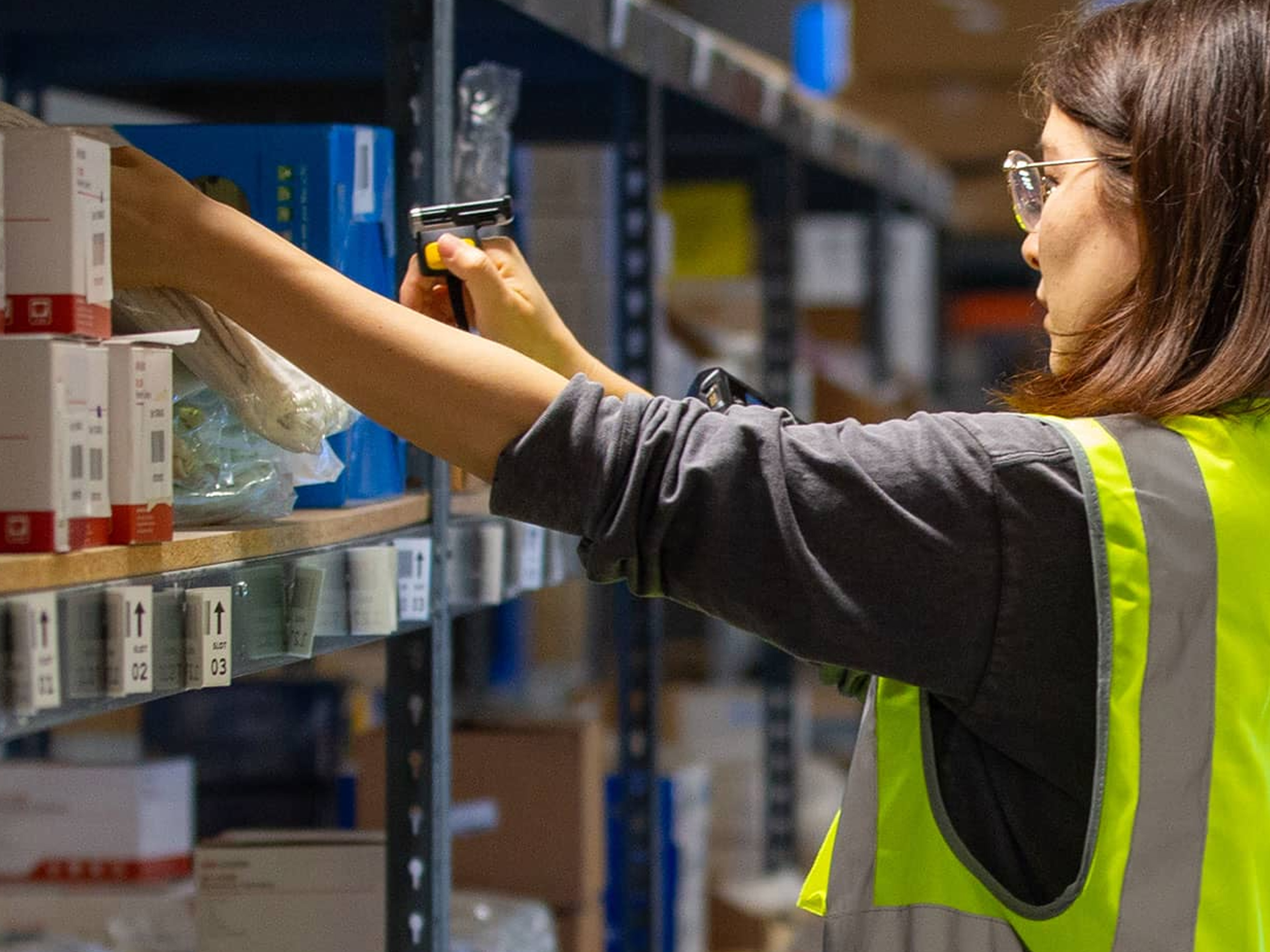 A person wearing a high-visibility vest scans an item on a warehouse shelf, seamlessly integrating into the Team Intelligence system, which ensures optimal coordination with autonomous mobile robots for enhanced efficiency.