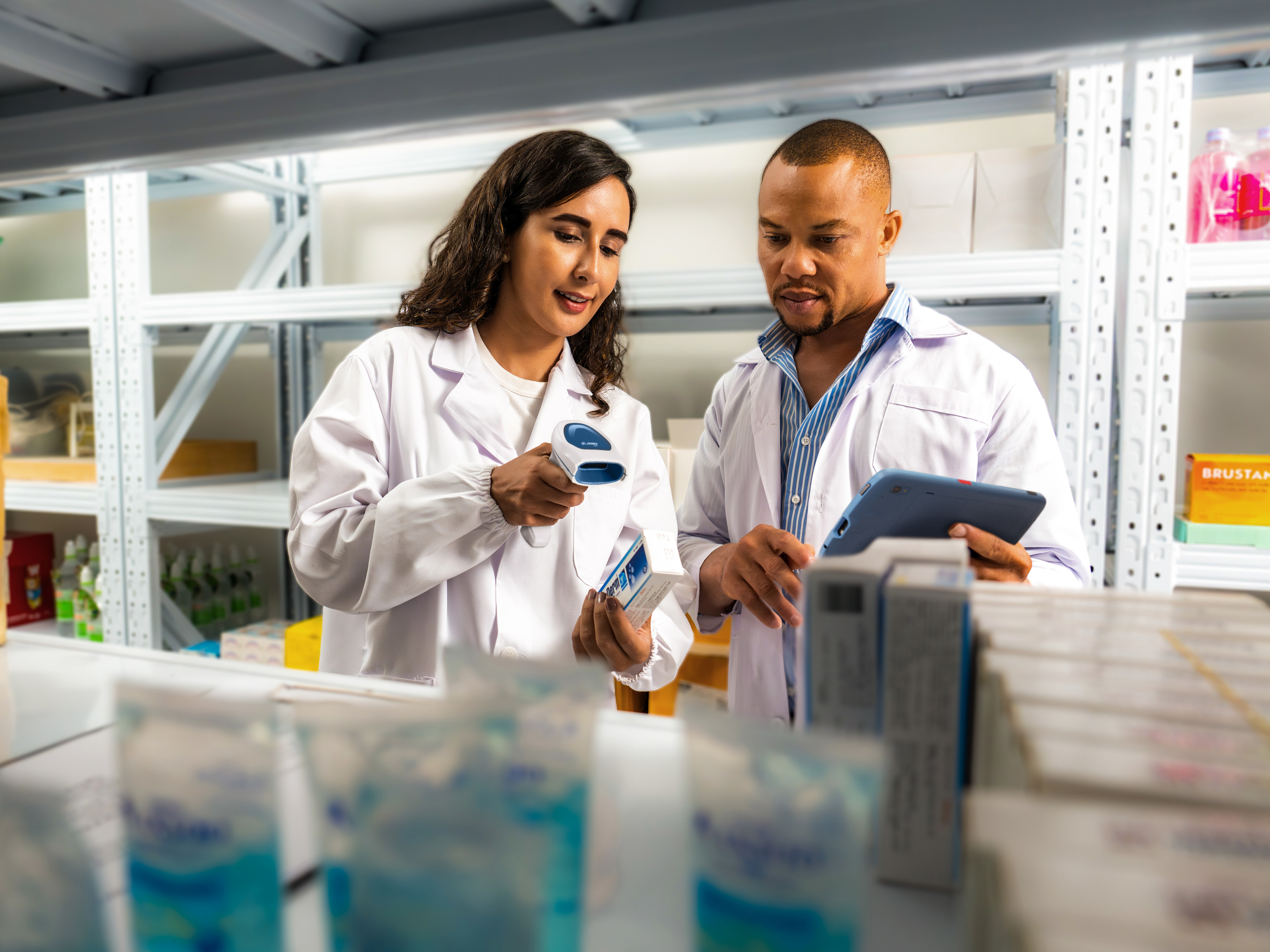 A medical professional uses a Zebra handheld barcode scanner to scan a label on a prescription medication while their colleague with a Zebra tablet watches.