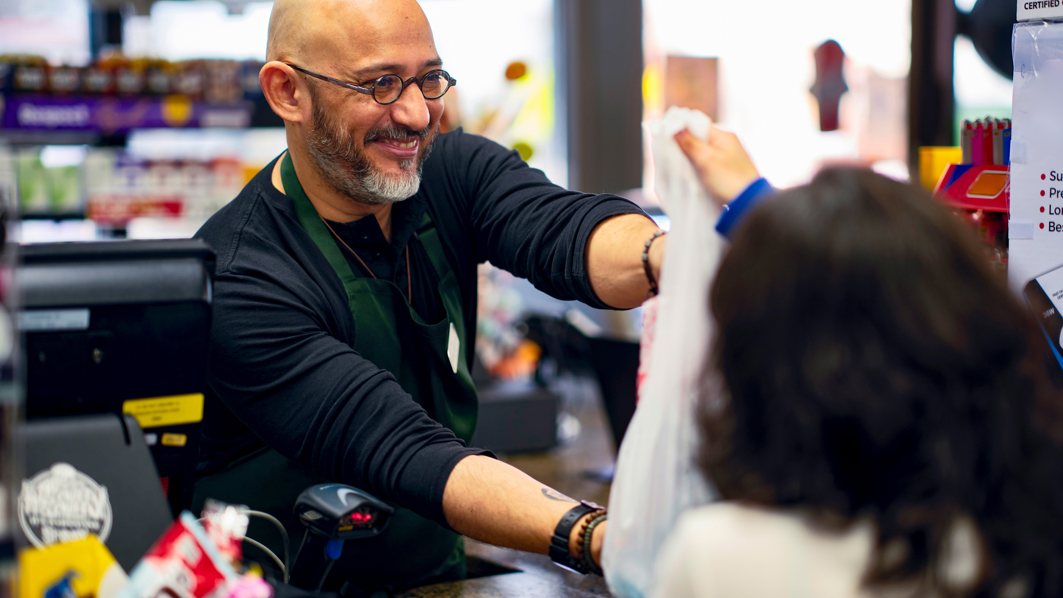 A cashier hands a plastic bag containing products to a customer at a checkout counter in a convenience store.