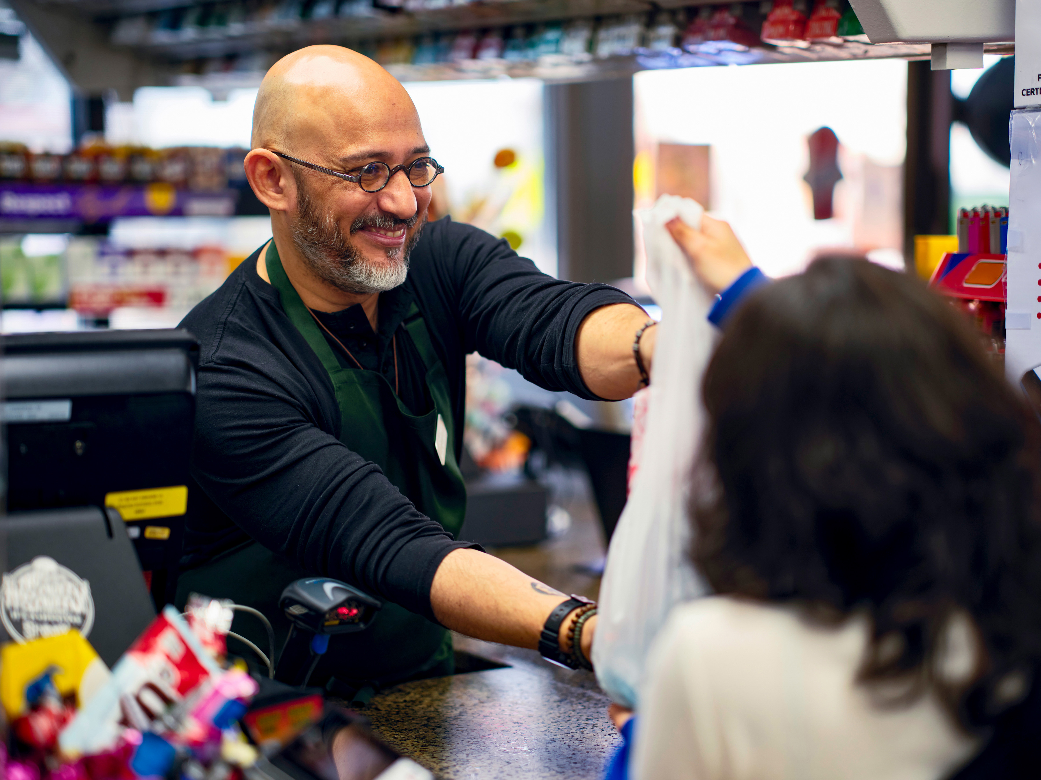 A cashier hands a plastic bag containing products to a customer at a checkout counter in a convenience store.