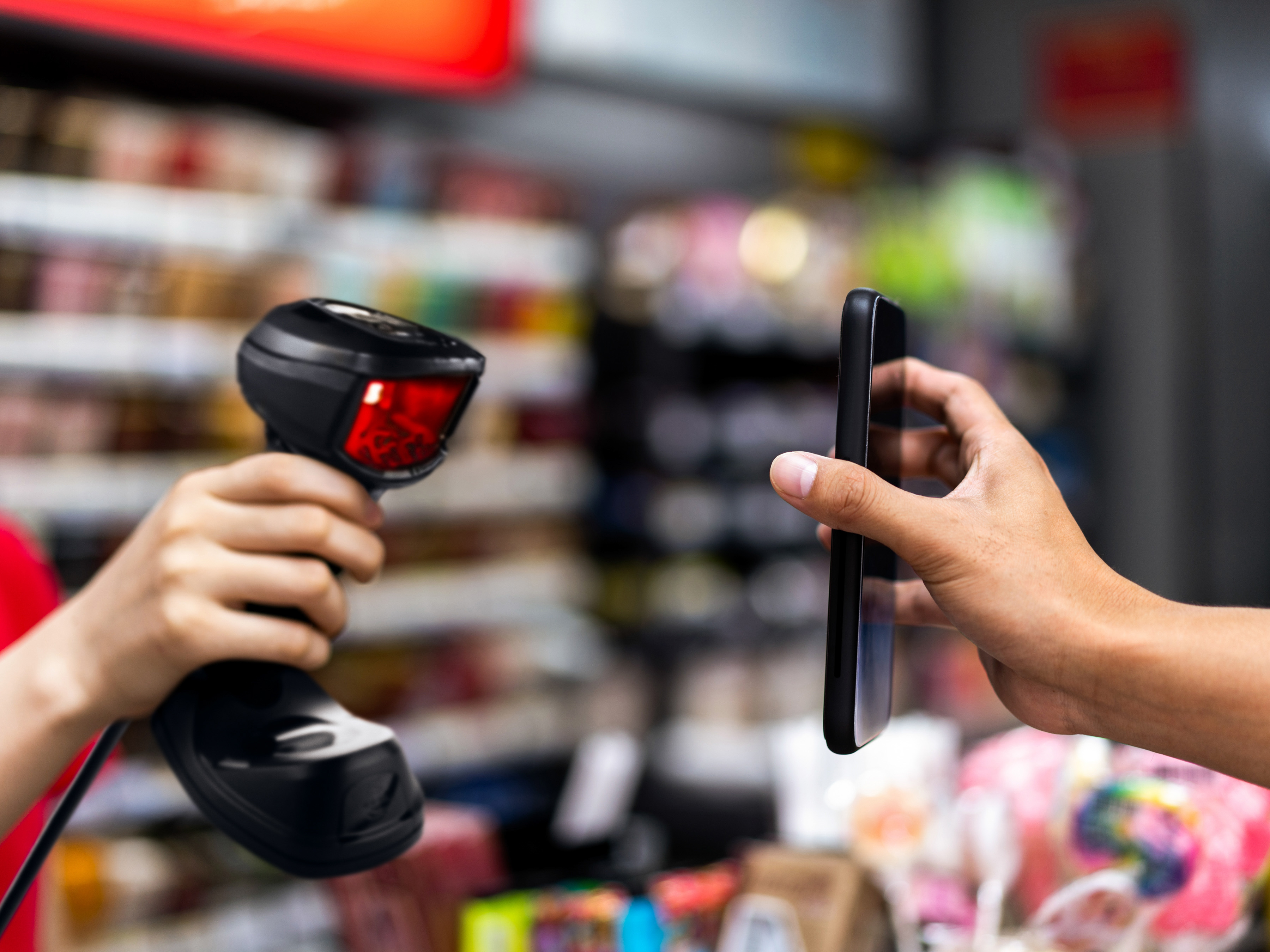 A retail employee using a handheld barcode scanner to scan a mobile application on a shopper's phone during checkout at a store.