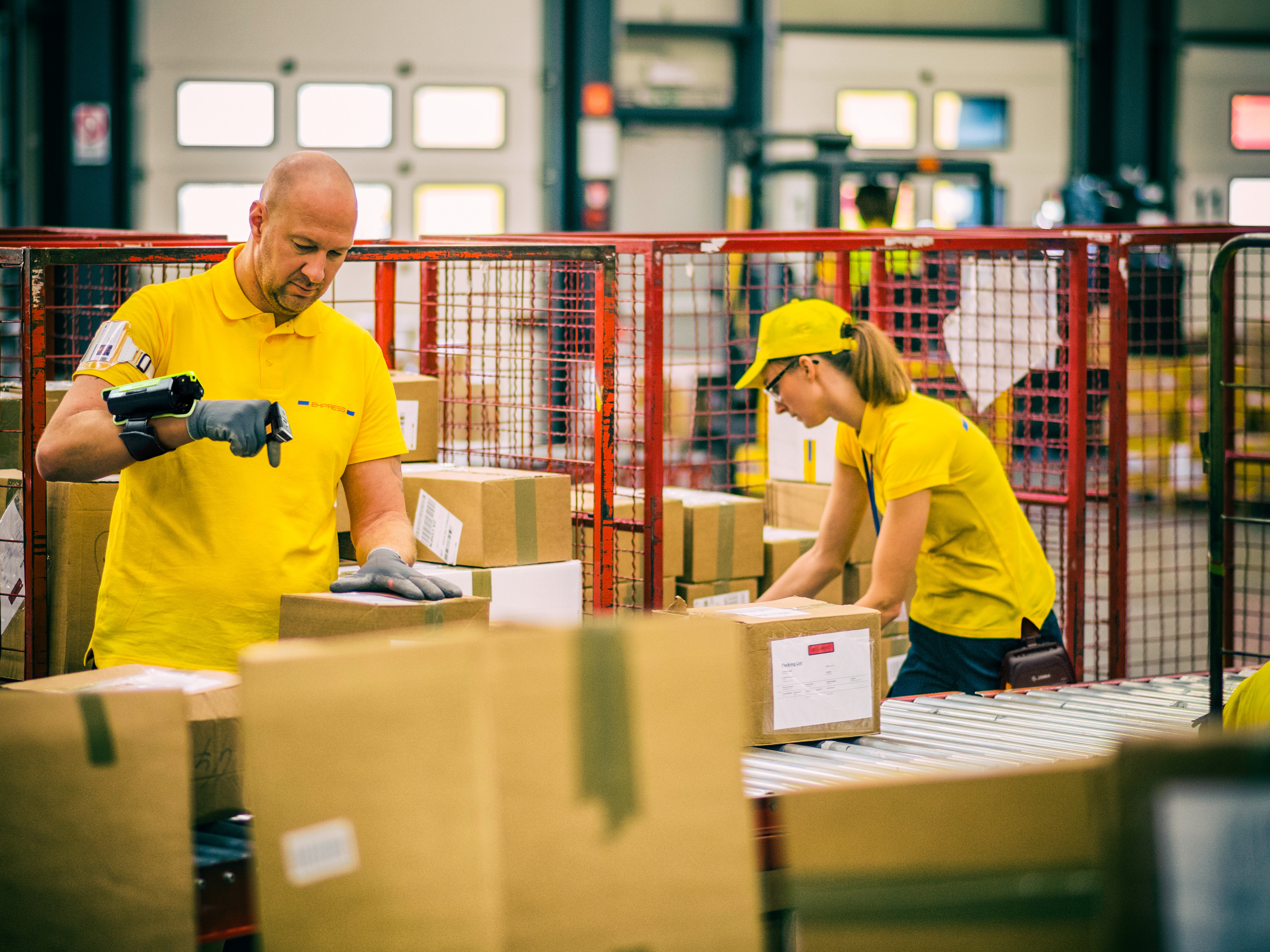 Two postal workers in yellow uniforms are sorting and scanning boxes using wearable scanners.
