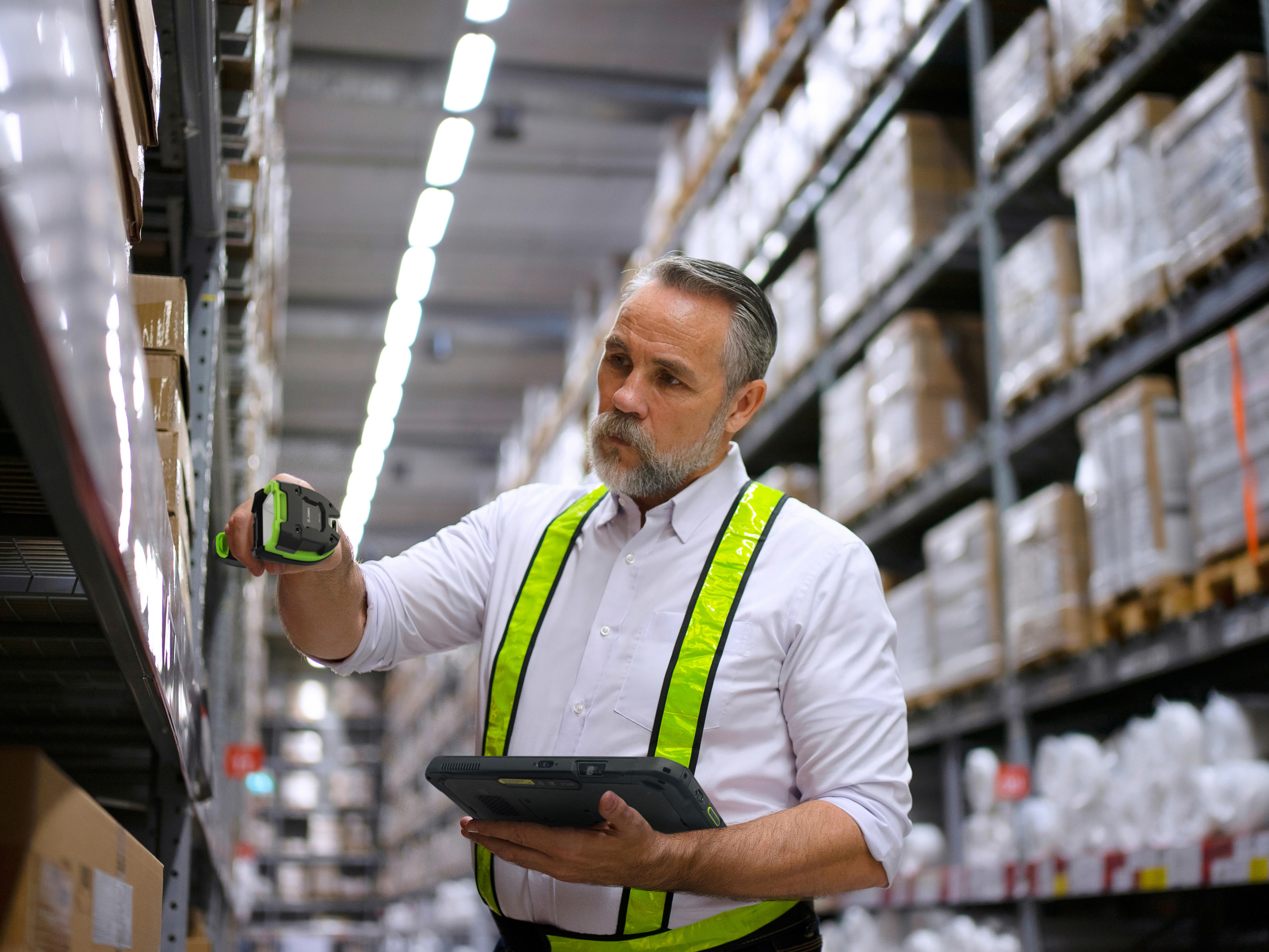 An employee using a handheld barcode scanner and a tablet to scan shelf stock inside of a warehouse.