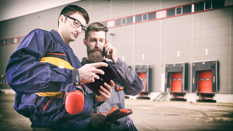 Two workers look at their rugged tablet screen as they stand outside dock doors